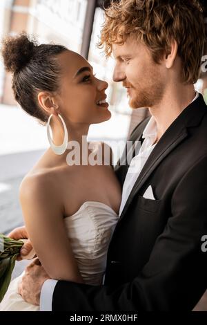 joyeux et élégant jeunes mariés interraciaux se regardant dans la rue, mariage dans un cadre urbain Banque D'Images