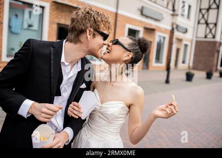 romance urbaine, mariage en plein air, jeunes mariés multiethniques dans des lunettes de soleil mangeant des frites ensemble Banque D'Images