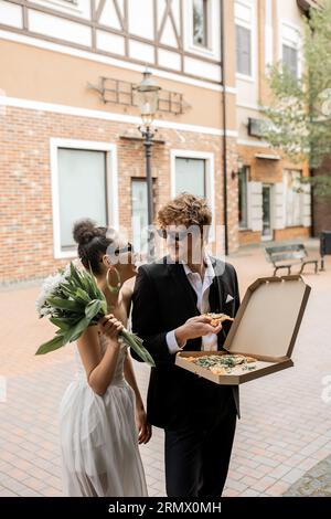 élégants jeunes mariés multiethniques avec des fleurs dans la pizza sur la rue urbaine, tenue de mariage, lunettes de soleil Banque D'Images