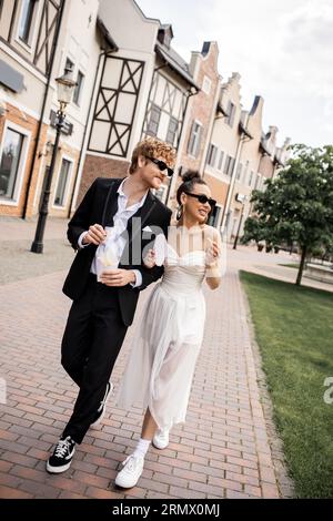 jeunes mariés multiethniques avec frites marchant dans la rue, lunettes de soleil, mariage en milieu urbain Banque D'Images