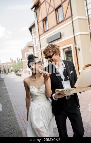 amour en ville, heureux jeunes mariés multiethniques dans des lunettes de soleil marchant avec pizza dans la rue Banque D'Images