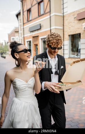 élégants jeunes mariés multiethniques dans des lunettes de soleil marchant avec pizza dans la ville européenne, mariage en plein air Banque D'Images