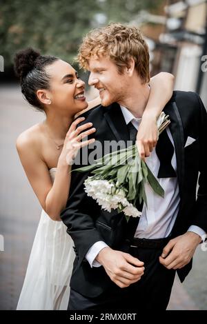 célébration de mariage en plein air, mariée afro-américaine excitée avec bouquet embrassant le marié roux Banque D'Images