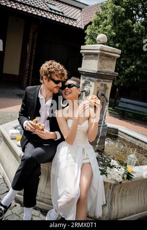 les jeunes mariés multiethniques aux lunettes de soleil grignotent avec des hamburgers et du jus d'orange près de la fontaine de la ville Banque D'Images