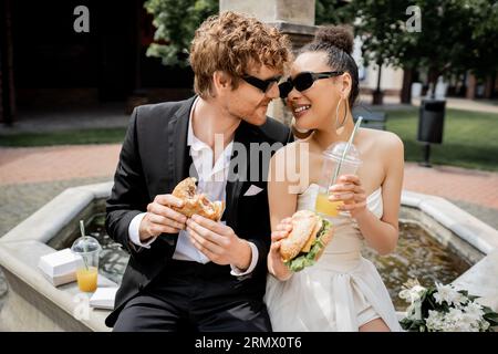 élégants jeunes mariés interraciaux en lunettes de soleil avec hamburgers et jus d'orange près de la fontaine de la ville Banque D'Images