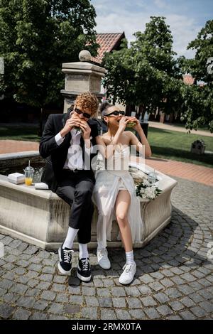 nouveaux mariés multiethniques dans des lunettes de soleil mangeant des hamburgers près de la fontaine, mariage dans la ville européenne Banque D'Images