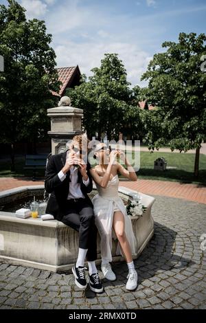élégants jeunes mariés interraciaux dans des lunettes de soleil mangeant des hamburgers près de la fontaine dans la ville européenne Banque D'Images