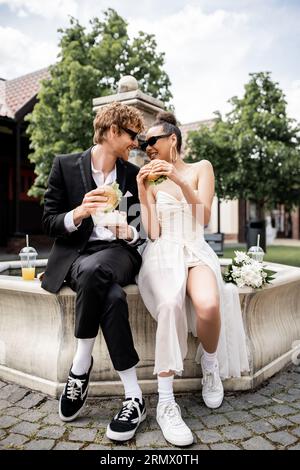 joyeux jeunes mariés interraciaux dans des lunettes de soleil tenant des hamburgers et se regardant près de la fontaine Banque D'Images