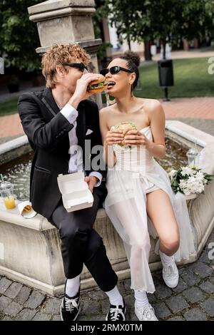 élégants jeunes mariés interraciaux dans les lunettes de soleil mangeant burger ensemble, rue de la ville, fontaine, plaisir Banque D'Images