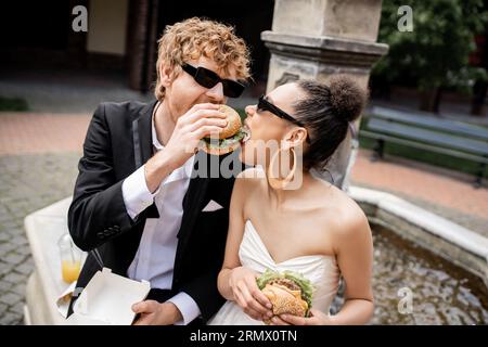 des jeunes mariés interraciaux portant des lunettes de soleil mordent s'amusant et mordent un hamburger ensemble près de la fontaine Banque D'Images