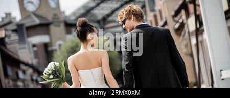 vue arrière des jeunes mariés interraciaux dans une tenue de mariage élégante sur la rue urbaine, bannière Banque D'Images