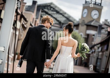 vue arrière de couple interracial élégant avec champagne et fleurs en ville, mariage en plein air Banque D'Images