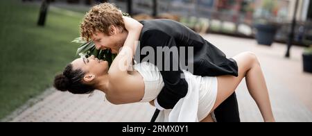 marié excité, roux et élégant embrassant heureuse mariée afro-américaine sur la rue urbaine, bannière Banque D'Images