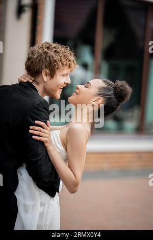 vue latérale du jeune marié embrassant la mariée afro-américaine joyeuse dans la ville, célébration en plein air Banque D'Images