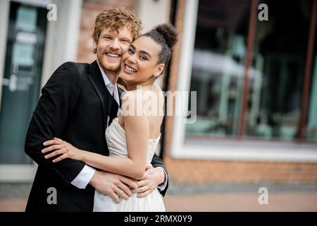 joyeux et élégant jeunes mariés multiethniques étreignant et regardant la caméra dans la rue, mariage en plein air Banque D'Images
