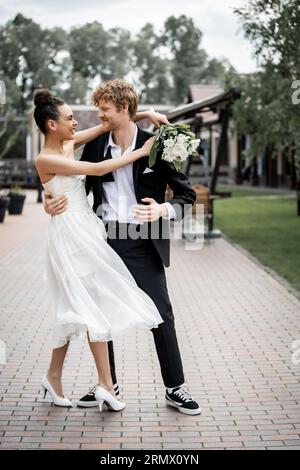 élégante mariée afro-américaine avec bouquet embrassant le marié souriant sur la rue, la joie, la romance urbaine Banque D'Images