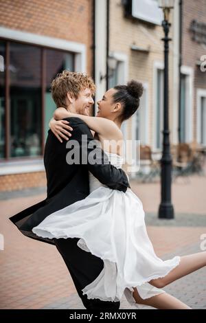 couple interracial en tenue de mariage embrassant sur la rue de la ville, bonheur, mariage en plein air, vue latérale Banque D'Images