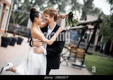 mariage en ville, mariée afro-américaine avec fleurs et heureux marié élégant embrassant dans la rue Banque D'Images