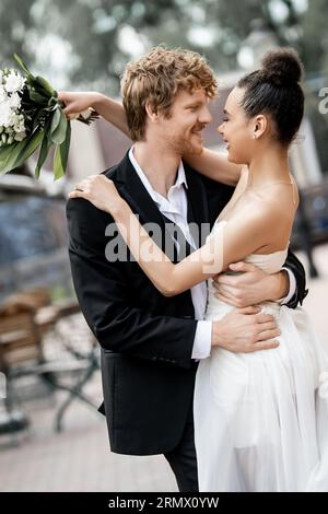 nouveaux mariés interraciaux dans une tenue élégante embrassant sur la rue, mariage en plein air, bannière Banque D'Images