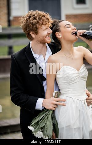 marié roux souriant serrant mariée afro-américaine buvant du champagne en bouteille, mariage en ville Banque D'Images