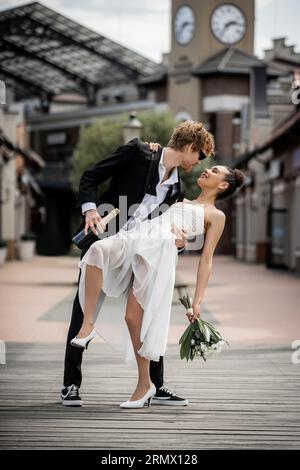 célébration de mariage, ville européenne, marié roux embrassant femme afro-américaine élégante dans la rue Banque D'Images