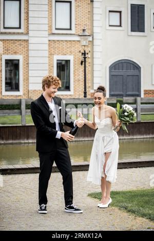 mariage en ville, marié ouvrant champagne près de mariée afro-américaine excitée avec des fleurs sur la rue Banque D'Images