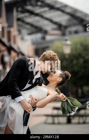 marié roux élégant embrassant la mariée afro-américaine avec bouquet sur la rue de la ville, mariage en plein air Banque D'Images