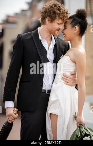 jeune homme rousse en costume élégant embrassant heureuse mariée afro-américaine, mariage en plein air Banque D'Images