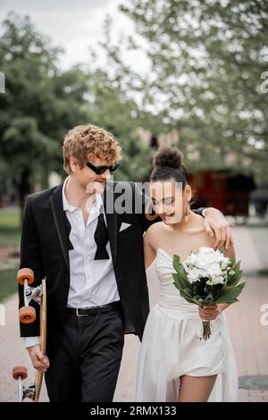 redhead groom dans des lunettes de soleil embrassant la mariée afro-américaine, marchant avec longboard et fleurs Banque D'Images