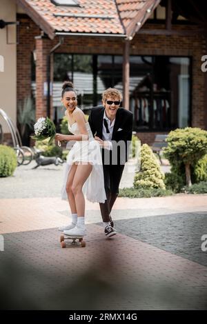 marié joyeux dans des lunettes de soleil soutenant l'élégante mariée afro-américaine chevauchant le longboard dans la rue Banque D'Images