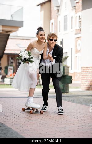 femme afro-américaine surjoyée dans la robe de mariée chevauchant longboard près de marié élégant sur la rue Banque D'Images