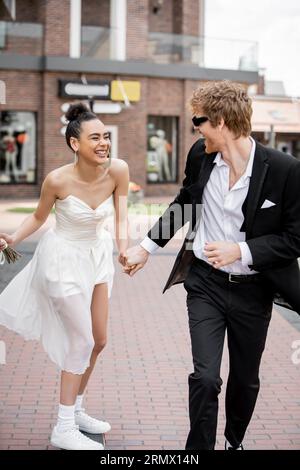 romance urbaine, élégants jeunes mariés interraciaux se tenant la main et courant dans la ville européenne Banque D'Images