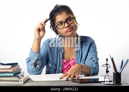 Une jolie jeune étudiante indienne en lunettes pensant tout en étudiant sur la table d'étude et fond blanc Banque D'Images