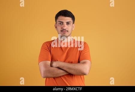 Portrait d'homme brésilien sérieux posant avec les bras croisés et regardant la caméra sur fond de studio orange Banque D'Images