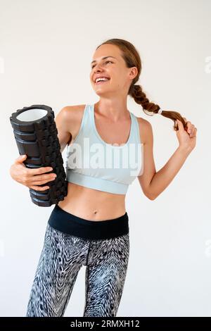 Athlète féminine heureuse de fitness posant devant le fond blanc avec le rouleau de mousse dans ses mains. Banque D'Images