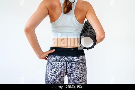 Vue arrière de la femme sportive debout devant le fond blanc dans le studio et tenant le rouleau de mousse dans la main. Banque D'Images
