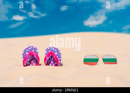 Lunettes de soleil avec des lunettes en forme de drapeau de la Bulgarie et tongs reposent sur le sable contre le ciel bleu. Concept de vacances d'été et de voyage en Bulgarie Banque D'Images