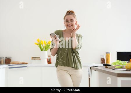 Heureuse jeune femme écoutant de la musique et dansant dans la cuisine légère Banque D'Images