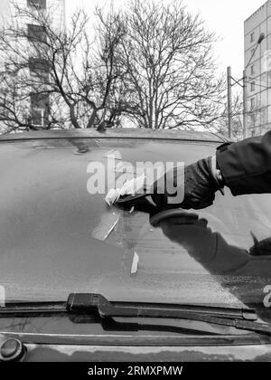 Nettoyage des vitres de voiture en hiver. L'homme gratte le cerceau avec un grattoir en plastique du pare-brise d'une voiture. Main dans un gant en cuir noir dans le froid. Banque D'Images