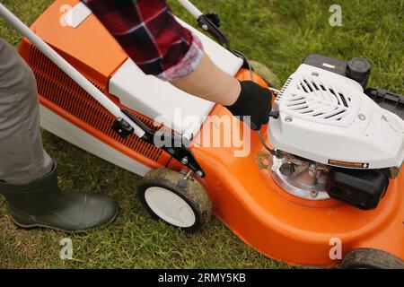 Un jardinier méconnaissable en gants de protection démarre le robot de tonte avant de couper de l'herbe verte dans son jardin. Homme avec tondeuse à gazon motorisé env Banque D'Images