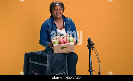 Courrier afro-américain transportant soigneusement le panier de fruits du sac de livraison de nourriture. Femme noire souriante livrant des fruits et légumes frais avec vélo comme moyen de transport. Banque D'Images