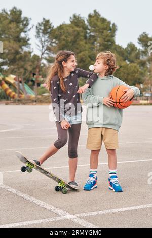 Un garçon et une jeune fille positif debout avec un skateboard et un basket-ball en ville et en train de souffler de la gomme à mâcher tout en s'amusant et en regardant les uns les autres Banque D'Images