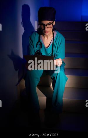 Corps entier de médecin féminin focalisé dans des lunettes avec stéthoscope assis sur les escaliers avec presse-papiers et lecture du rapport médical dans la chambre avec la lumière néon Banque D'Images