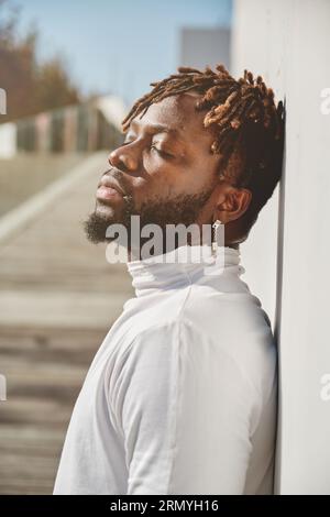 Portrait vue de côté d'un homme barbu afro-américain pensif avec des dreadlocks et boucle d'oreille debout près du mur avec les yeux fermés Banque D'Images