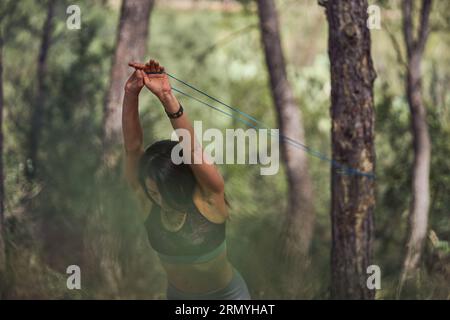 Femme en bonne santé levant les mains au-dessus de la tête tout en tirant la bande de résistance attachée sur le tronc d'arbre pendant l'entraînement actif dans les bois Banque D'Images