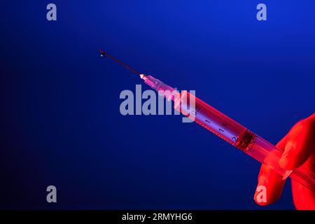 Seringue médicale avec aiguille pleine de vaccin pour COVID19 dans la main du médecin anonyme sur fond bleu en studio avec lumière rouge Banque D'Images