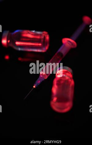 Mise au point sélective de la seringue médicale complète et des flacons brouillés avec le vaccin contre le coronavirus sur fond noir dans un éclairage au néon rouge Banque D'Images