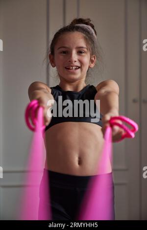 Gymnaste de pré-teen souriant en vêtements de sport noir et avec chignon de cheveux s'exerçant avec la corde à sauter rose dans la chambre et regardant la caméra Banque D'Images