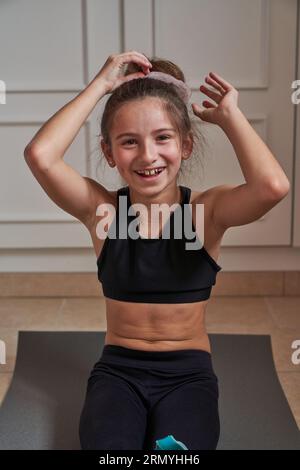 Souriante fille préadolescente en vêtements de sport reposant sur le tapis gris et regardant la caméra tout en faisant chignon de cheveux pendant la pause dans l'entraînement de gymnastique à la maison Banque D'Images
