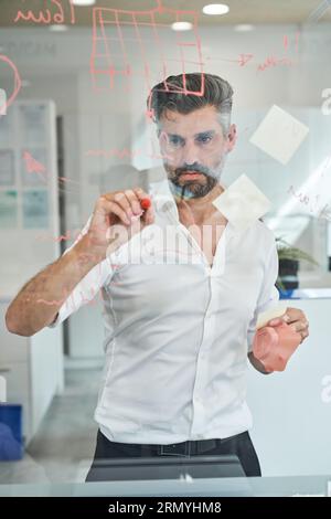Scientifique masculin hispanique concentré non rasé dessinant un croquis de masque médical à partir du coronavirus et calculant des indicateurs tout en travaillant en laboratoire Banque D'Images